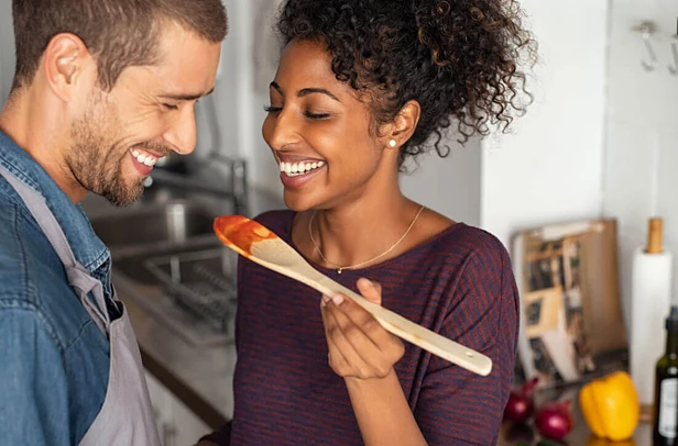 Two people smiling in a kitchen; one holds a wooden spoon with red sauce while facing the other.