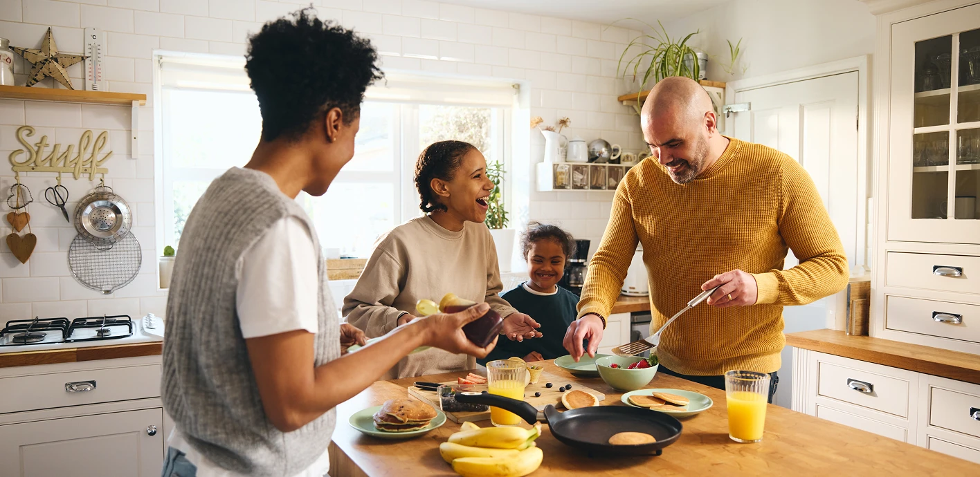 Three adults and a child laugh while preparing breakfast at a kitchen island with pancakes, fruit, and orange juice.