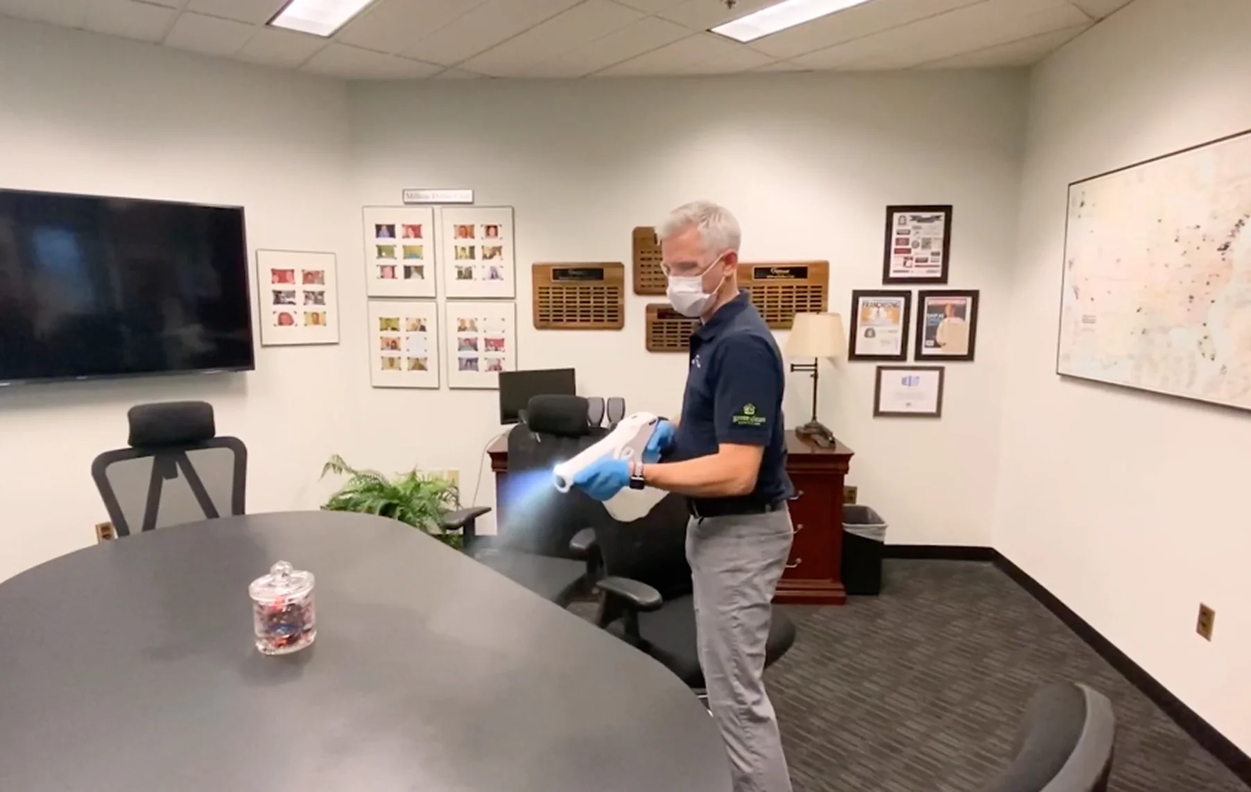Man wearing a face mask and gloves disinfects a conference room table with a handheld sprayer.